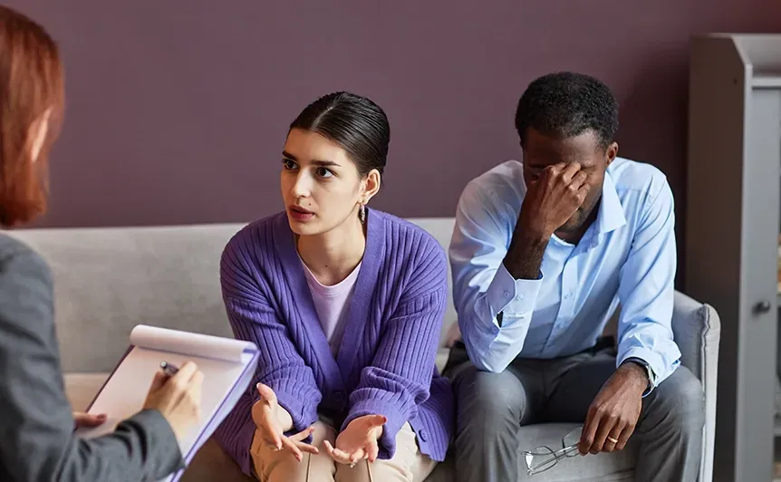 Two people sit on a couch during a counseling session, one looking concerned and the other covering his face.
