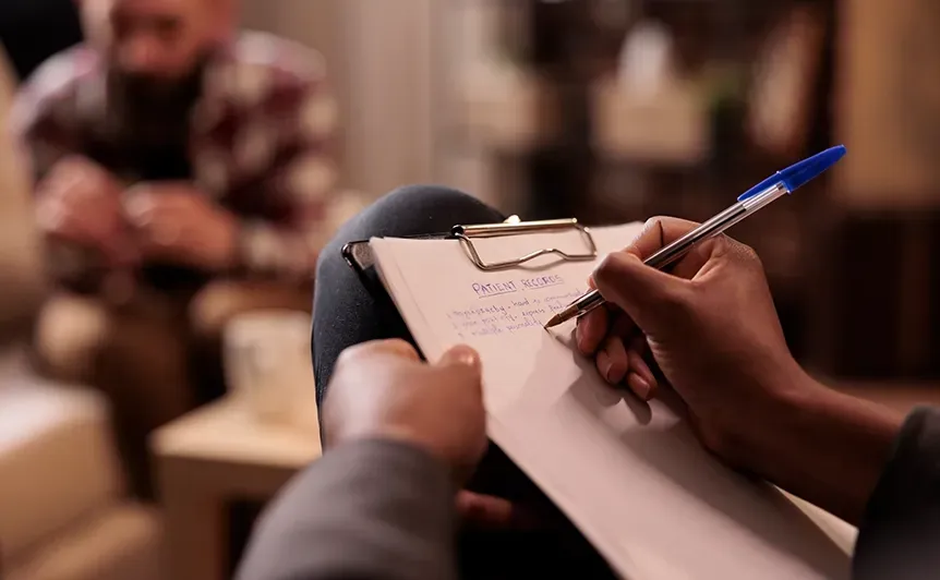Close-up of hands writing on a clipboard with a blue pen, with a blurred audience in the background