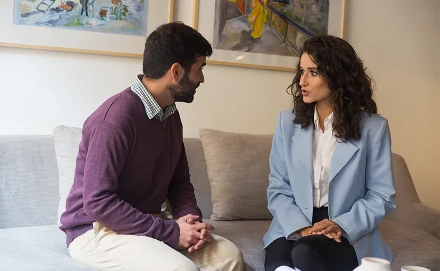 Two people sit on a couch talking in a bright living room.