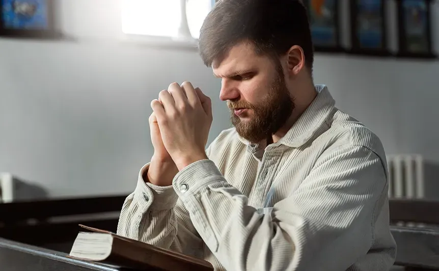 Man praying at a table with an open book in a softly lit room