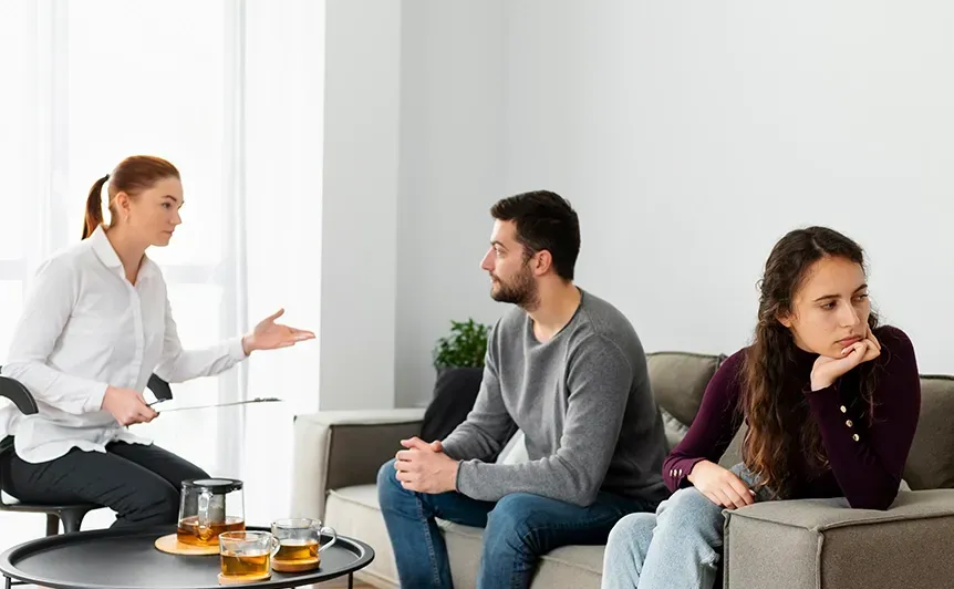 Woman speaking to two seated people in a bright office lounge, with coffee cups on a table.