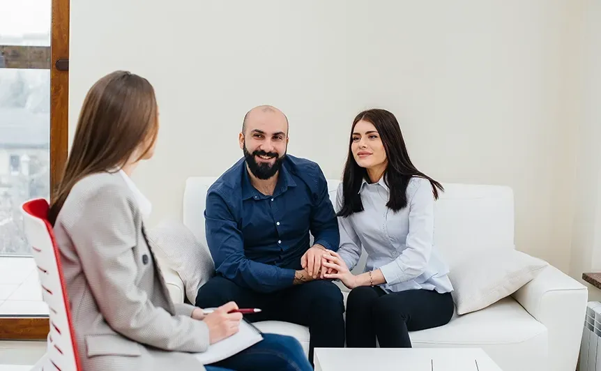 Three people talking in a bright living room, seated on a white couch with one holding a notebook.