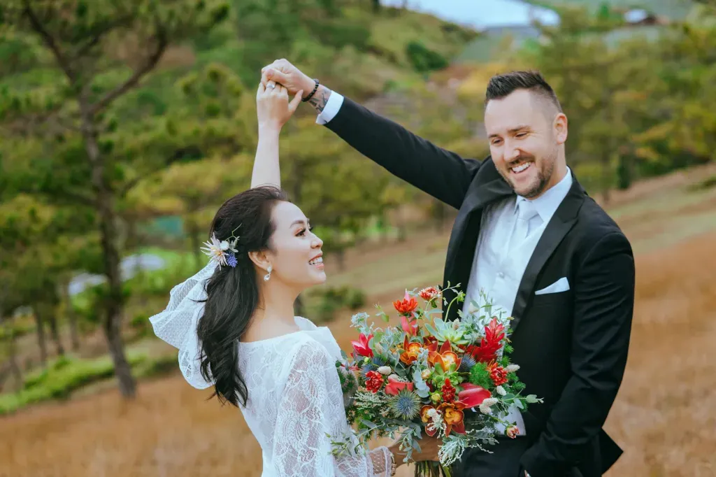 Couple dancing outdoors, bride in white lace dress and groom in black suit holding a colorful bouquet