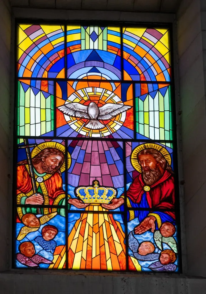 Stained-glass church window with a dove, angels, and a radiant central light in blue, gold, and red.