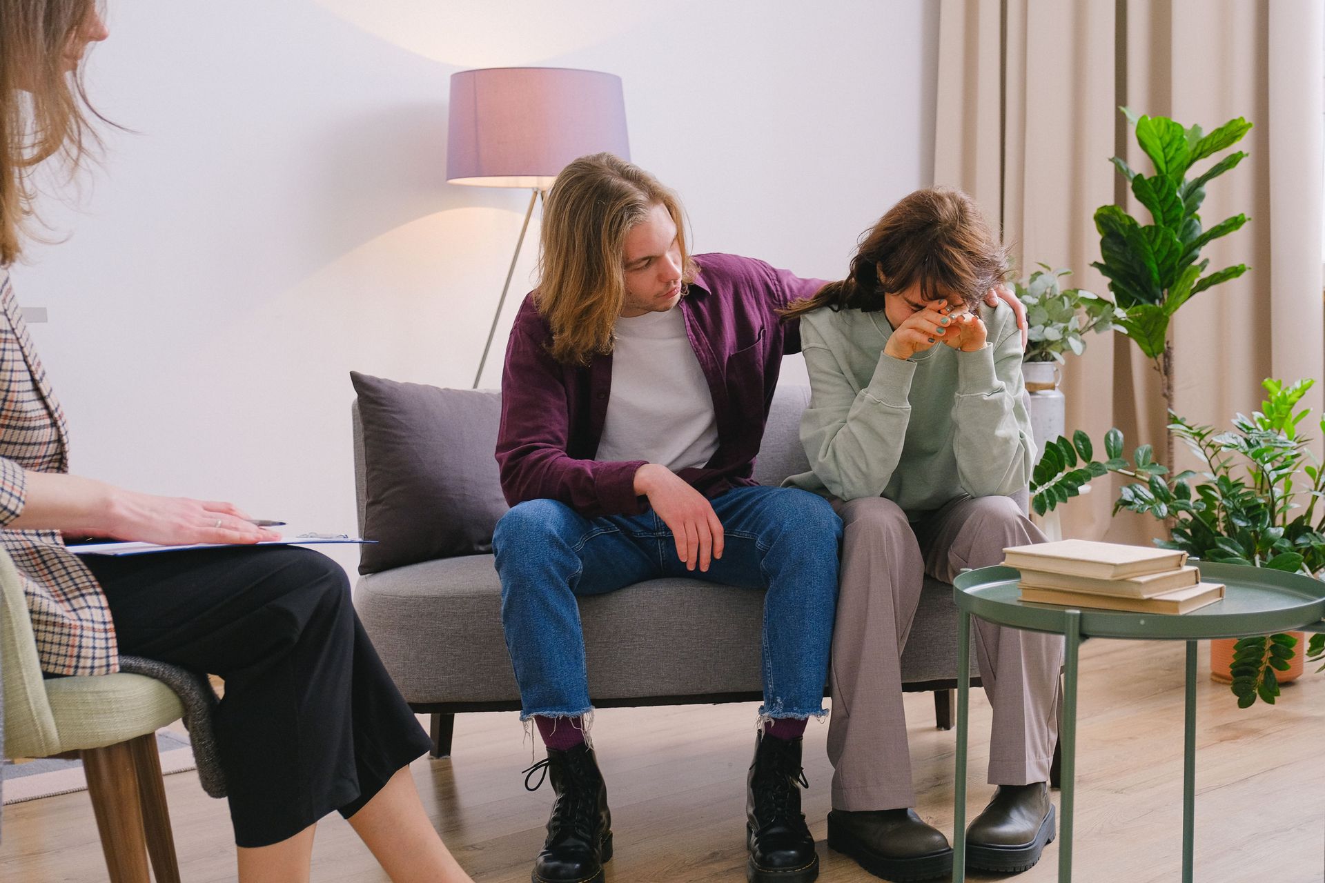 Two people sit on a couch in a living room, one consoling the other who looks upset.