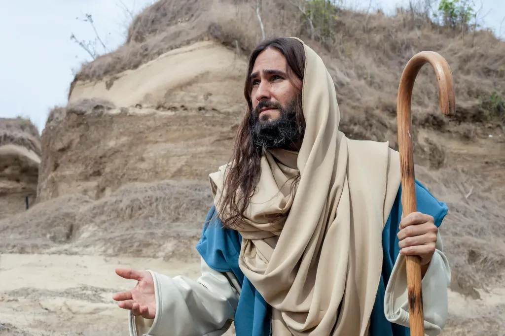 Bearded figure in beige robe and blue cloak holding a staff in a rocky desert landscape