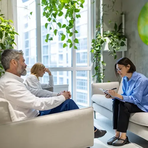 Three people in a bright office lounge discussing notes on a clipboard and laptop.
