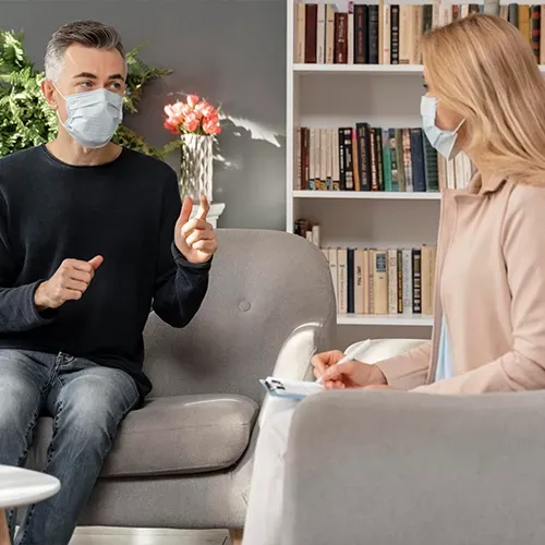 Two masked people talking in a bright living room, seated apart on sofas.