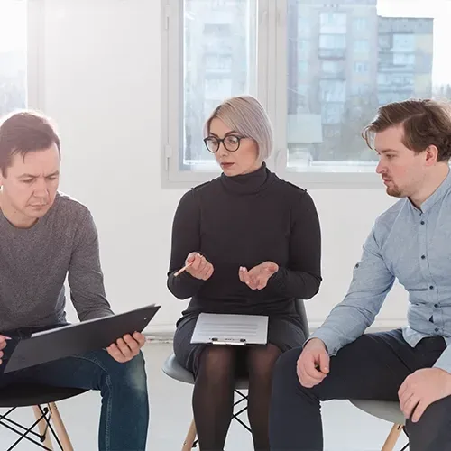 Three coworkers in a bright office discuss a tablet and notes during a meeting.