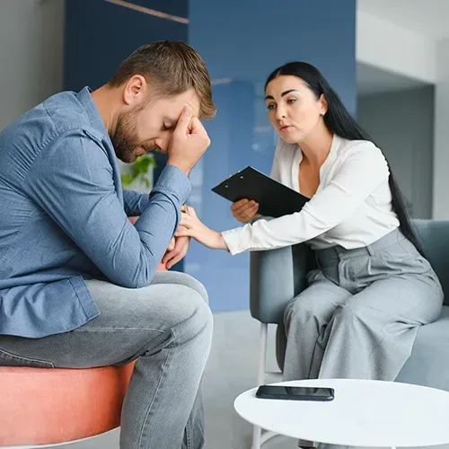 Man in blue shirt sits with head in hand as a woman in white talks with a clipboard in a counseling room