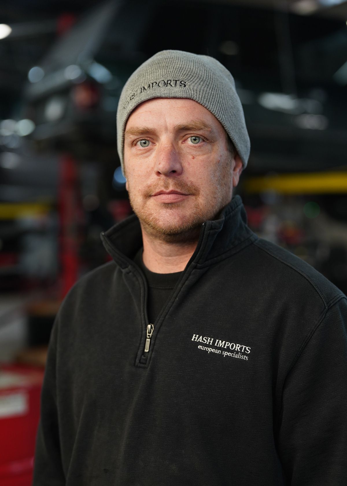A person wearing a grey beanie and a black quarter-zip jacket, standing in a workshop setting with machinery.