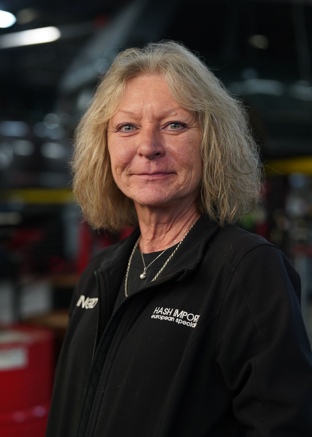 A person with shoulder-length blonde hair, wearing a black jacket with a logo, stands in a blurred workshop setting.