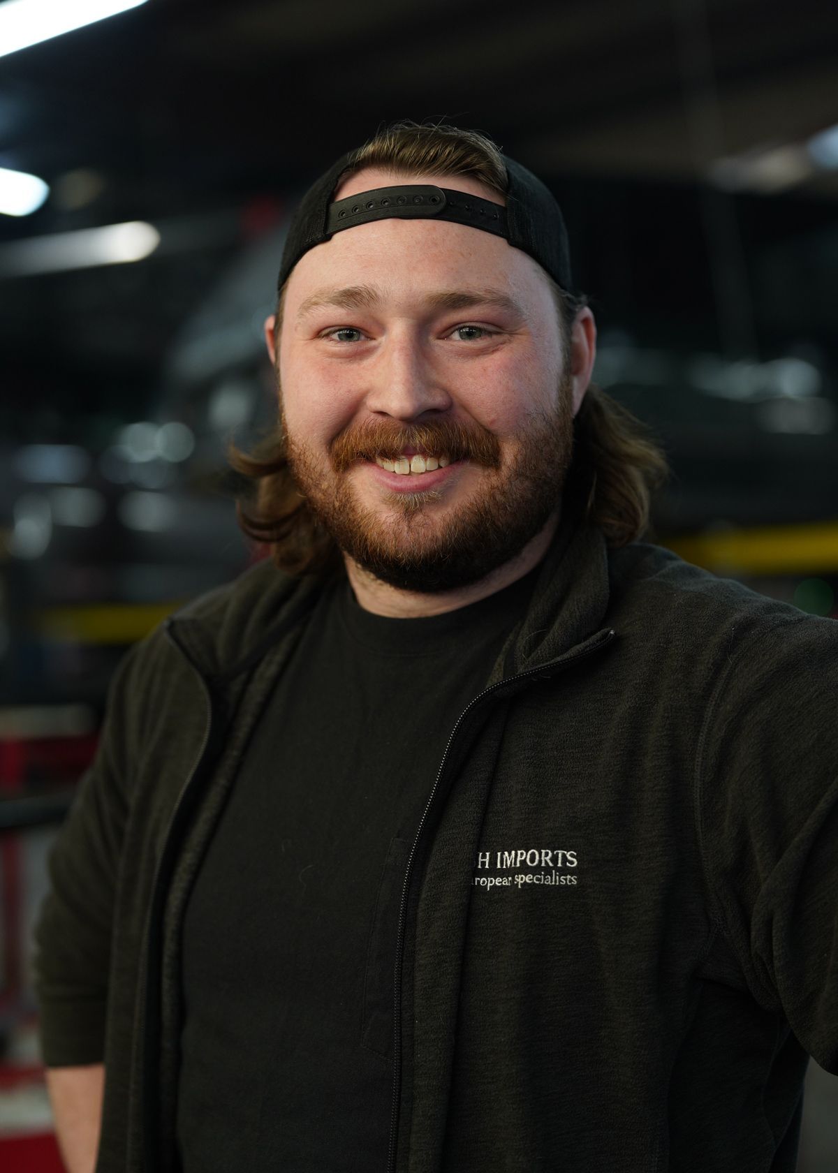 Smiling person with a beard and a backwards baseball cap wearing a dark zip-up jacket in a workshop setting.