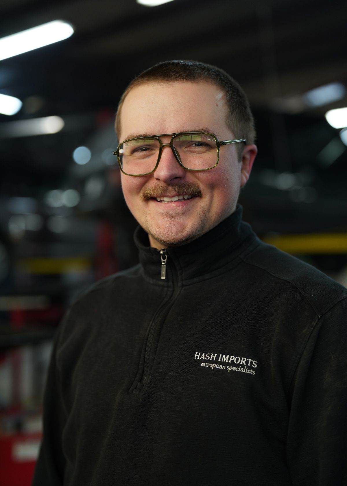 A person wearing wire-rimmed glasses and a black quarter-zip fleece, smiling in an industrial workshop setting.