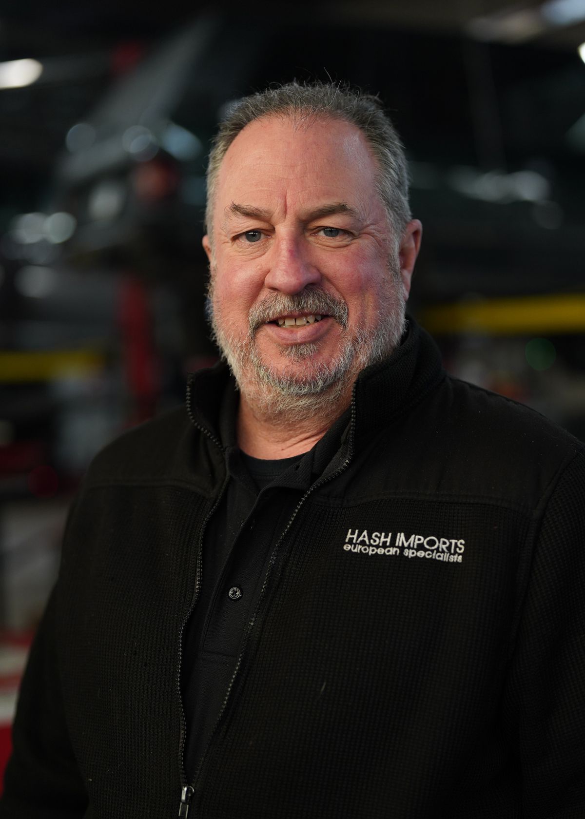 A man with a graying beard, wearing a black zip-up jacket with a logo, standing in a workshop setting.