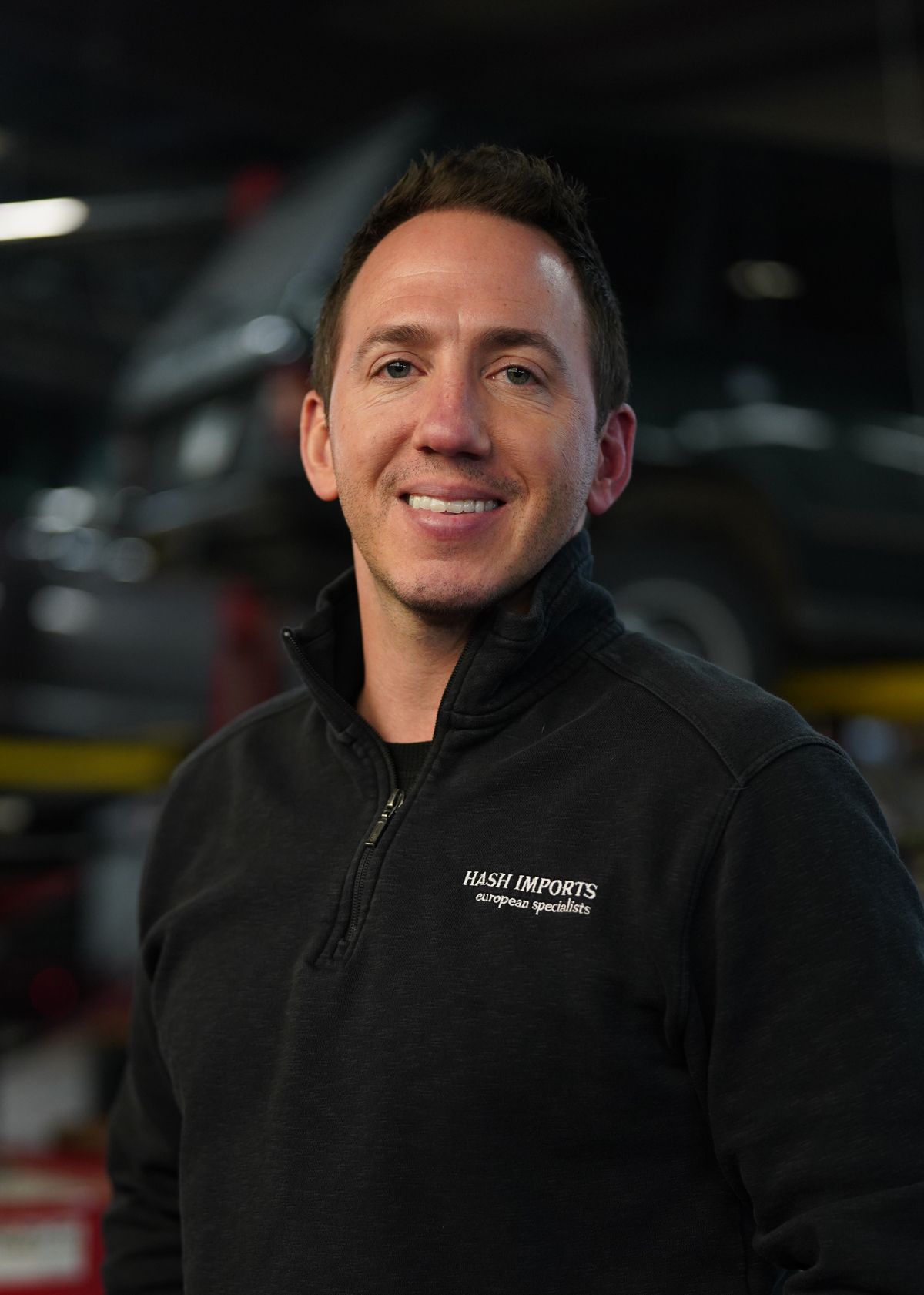A man smiling in a black quarter-zip fleece, standing in an automotive shop with a vehicle lift in the blurred background.