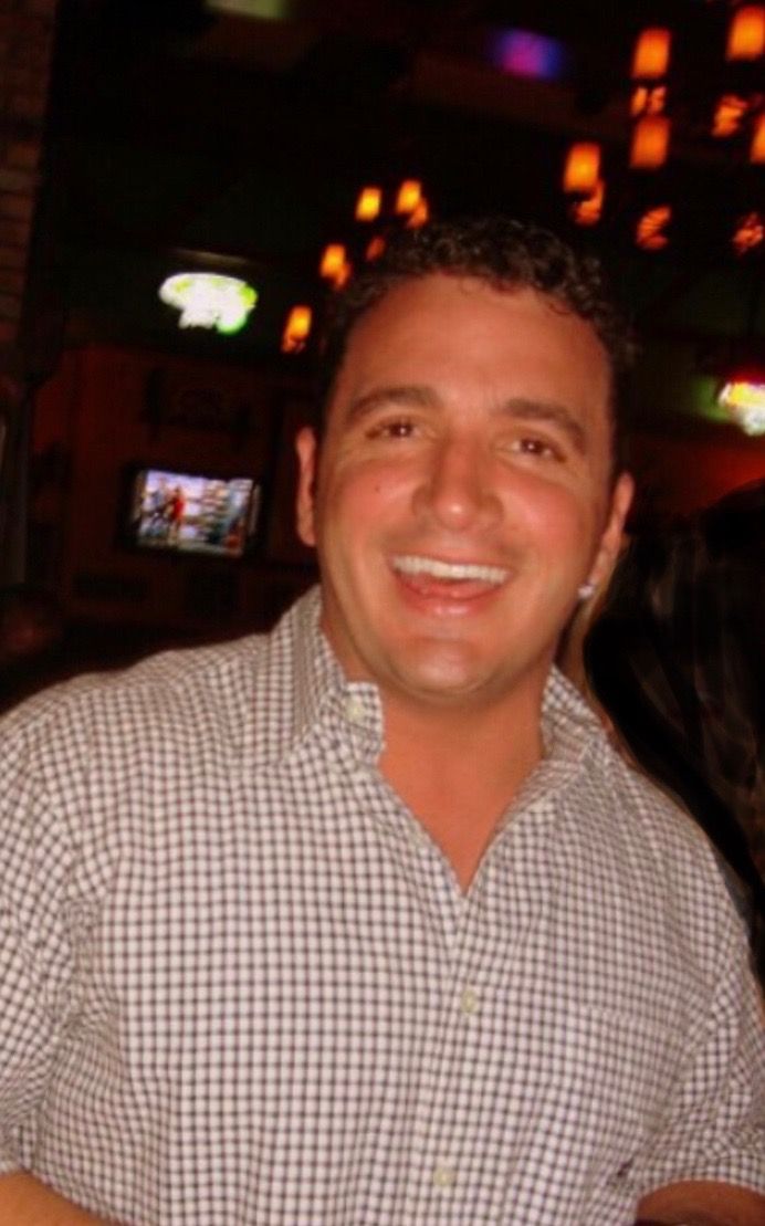 A smiling person wearing a light-colored, checkered button-down shirt, standing in a dimly lit indoor setting.