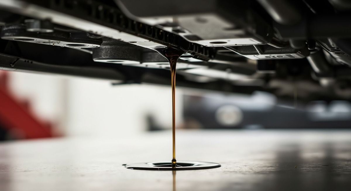 Used motor oil draining from the underside of a vehicle into a dark pool on a workshop floor.