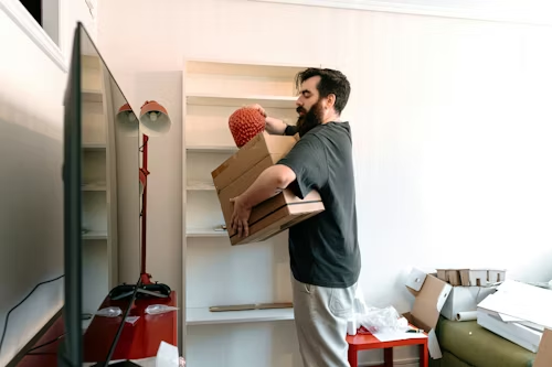 Man carrying a cardboard box into a room with a TV, red vacuum, and scattered moving boxes