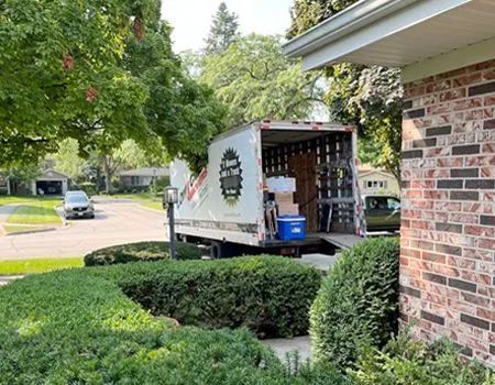 Moving truck parked in front of a brick house, boxes visible in the open trailer, shrubs in the foreground, sunny day.