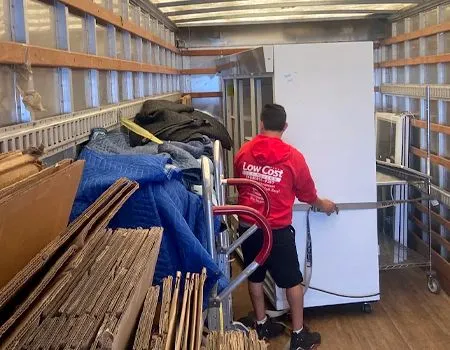 Mover in red shirt loading a white refrigerator into a moving truck packed with furniture and boxes