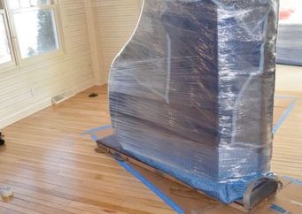 Piano wrapped in blue plastic on a dolly inside a room with wood floors.