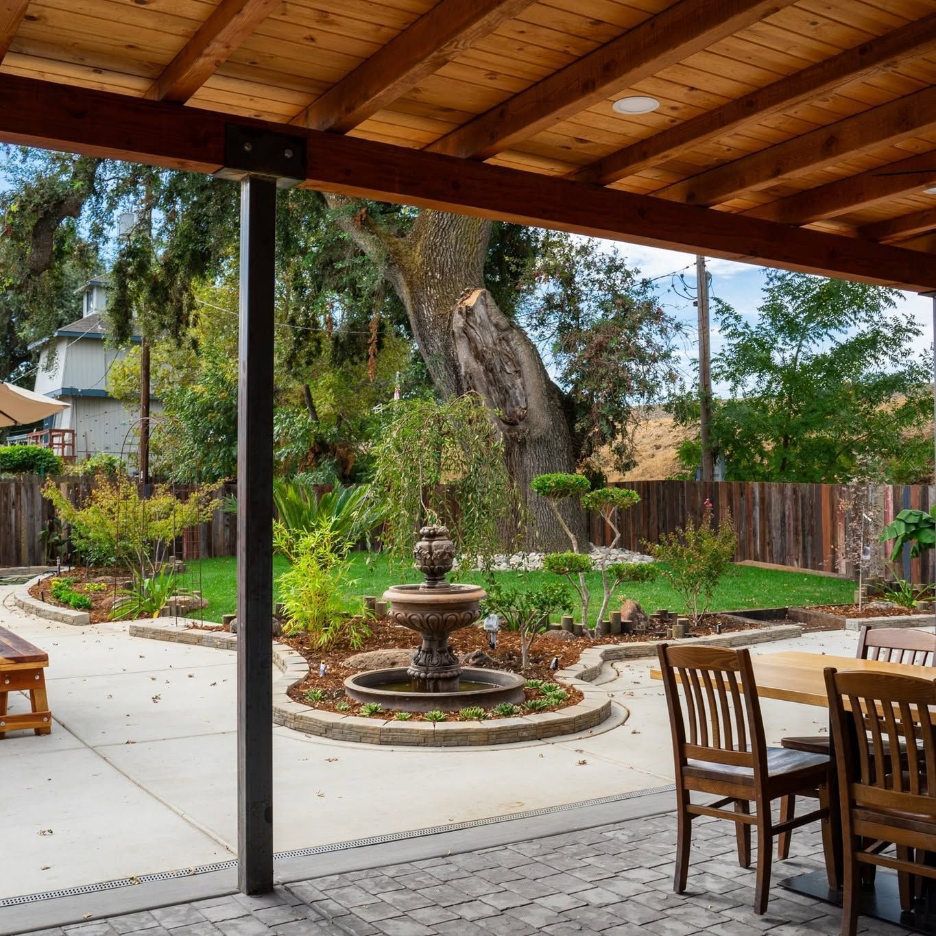 Covered patio overlooking a landscaped yard with a central fountain, wooden dining table, and chairs