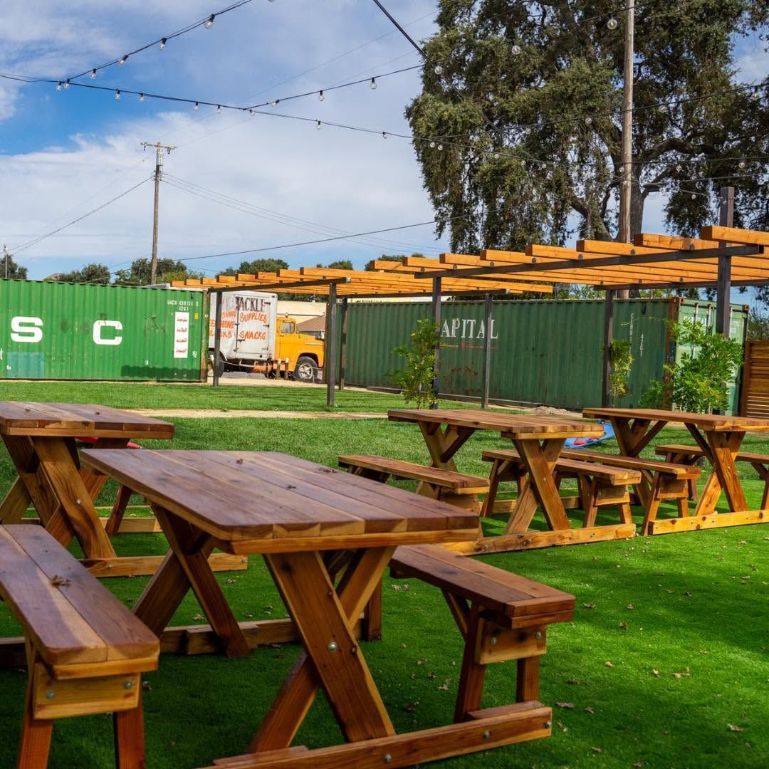 Wooden picnic tables on green grass beside shipping containers under string lights on a sunny day