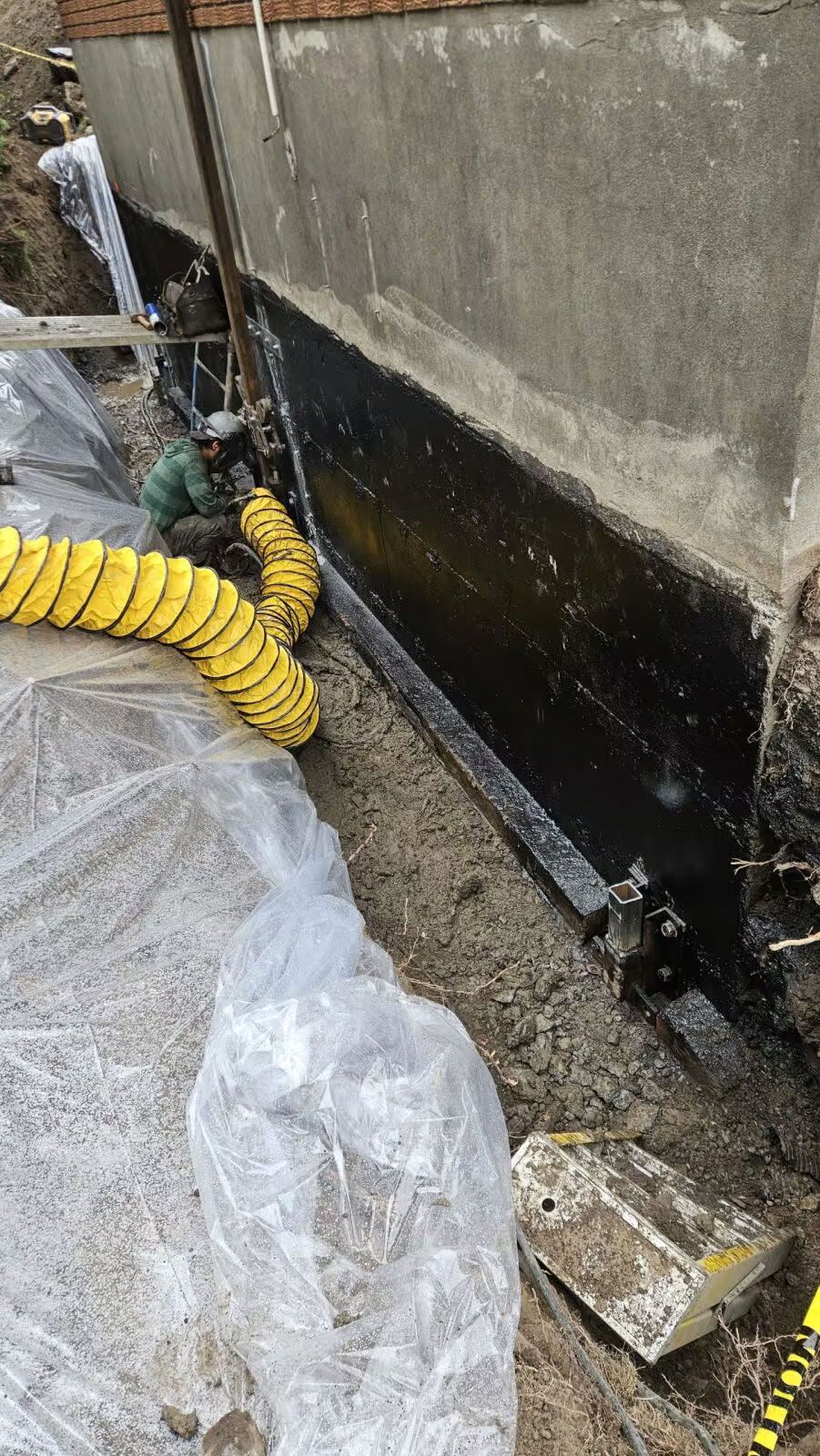 Travaux d'étanchéité sur un mur en béton, avec revêtement noir, tuyau jaune et bâche en plastique au sol.