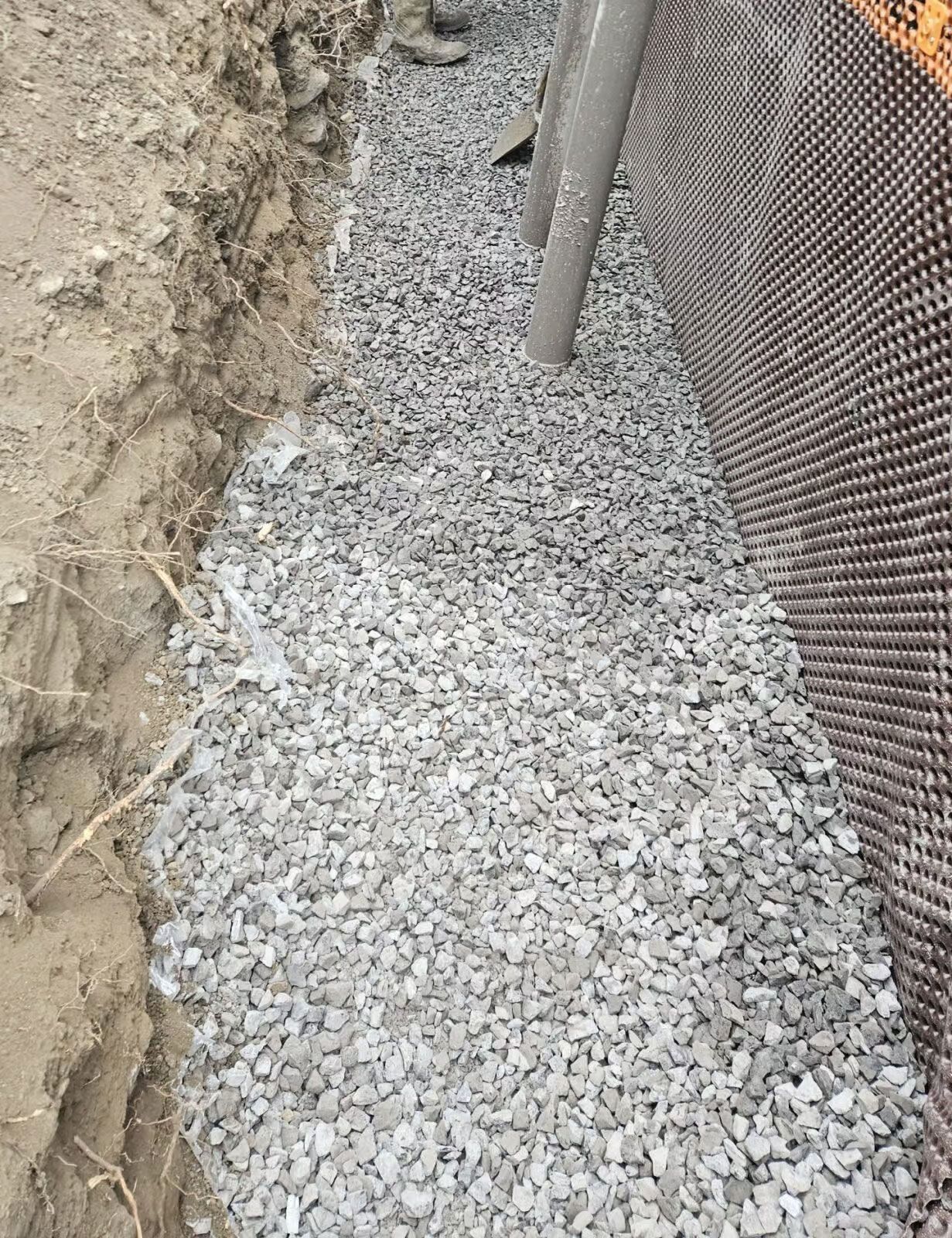 Tranchée remplie de gravier le long d'un mur de fondation avec membrane de drainage et tuyau. Terre à gauche.