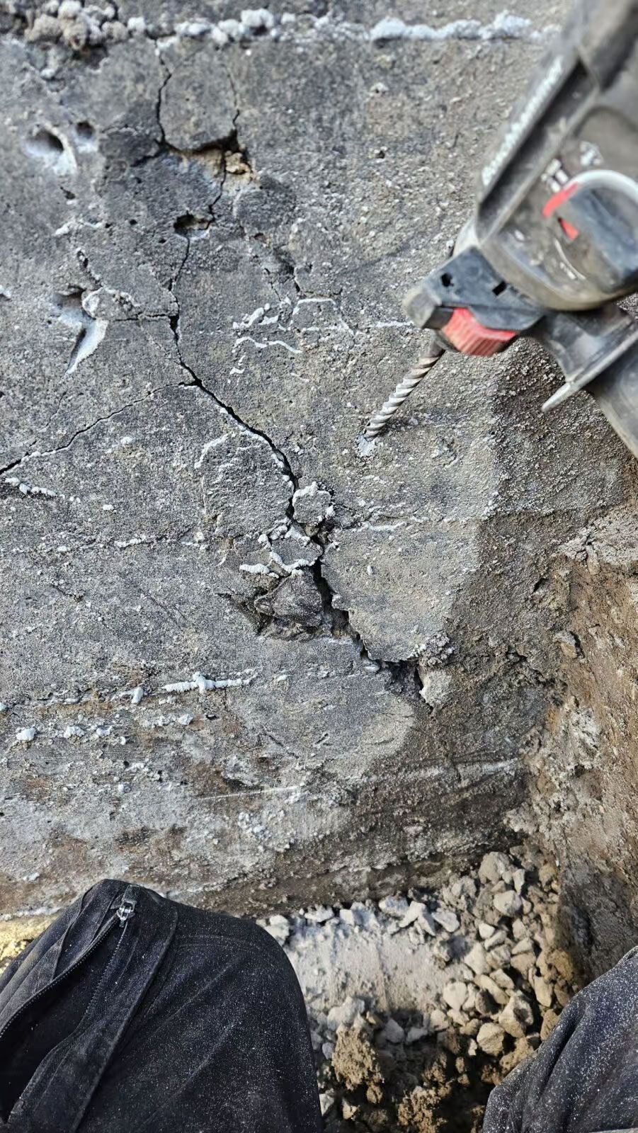 Gros plan sur du béton fissuré percé à l'aide d'un outil électrique ; la jambe de l'ouvrier est visible.