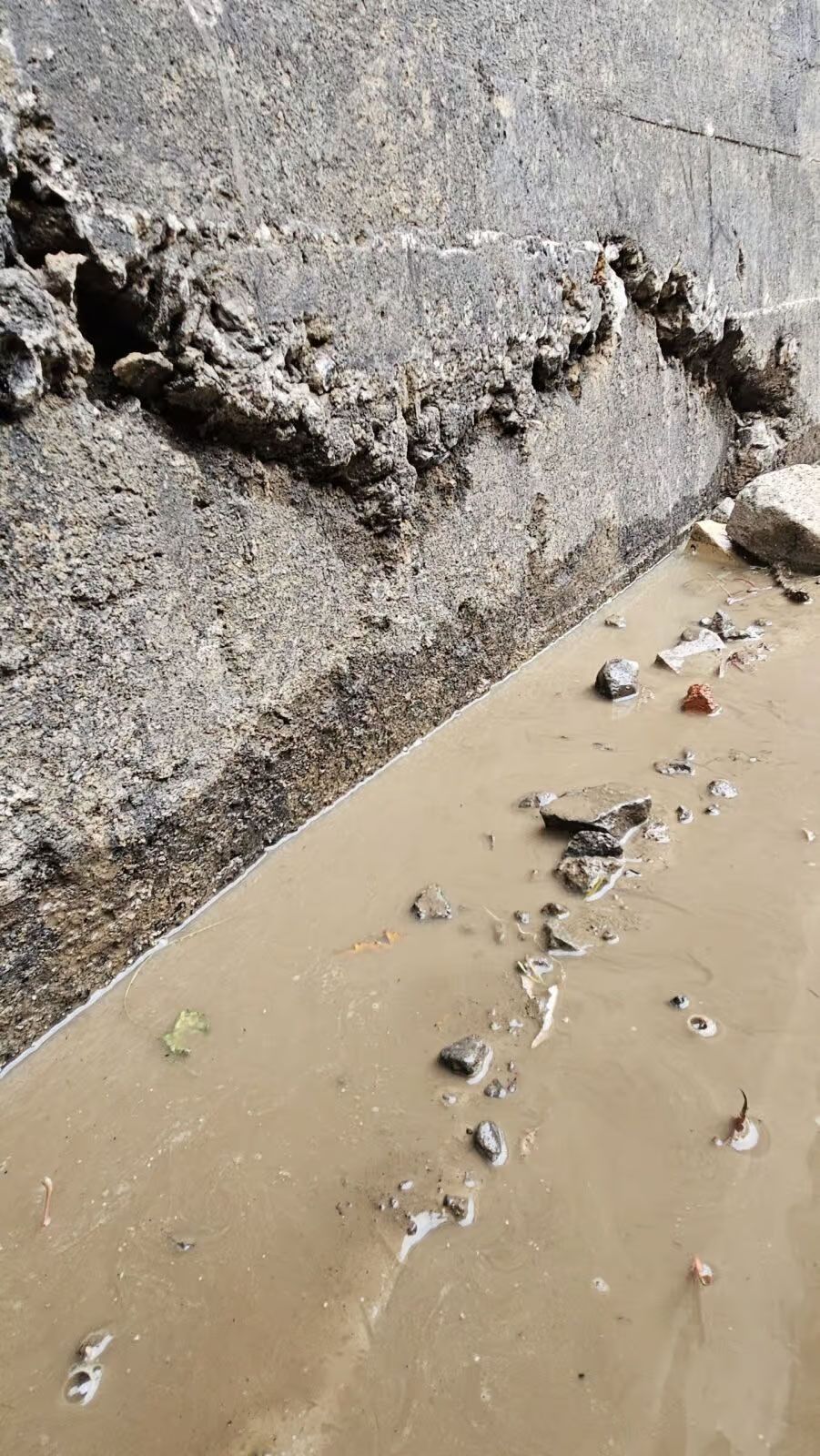Mur en béton endommagé, partiellement submergé par des eaux troubles. Débris et fissures visibles.