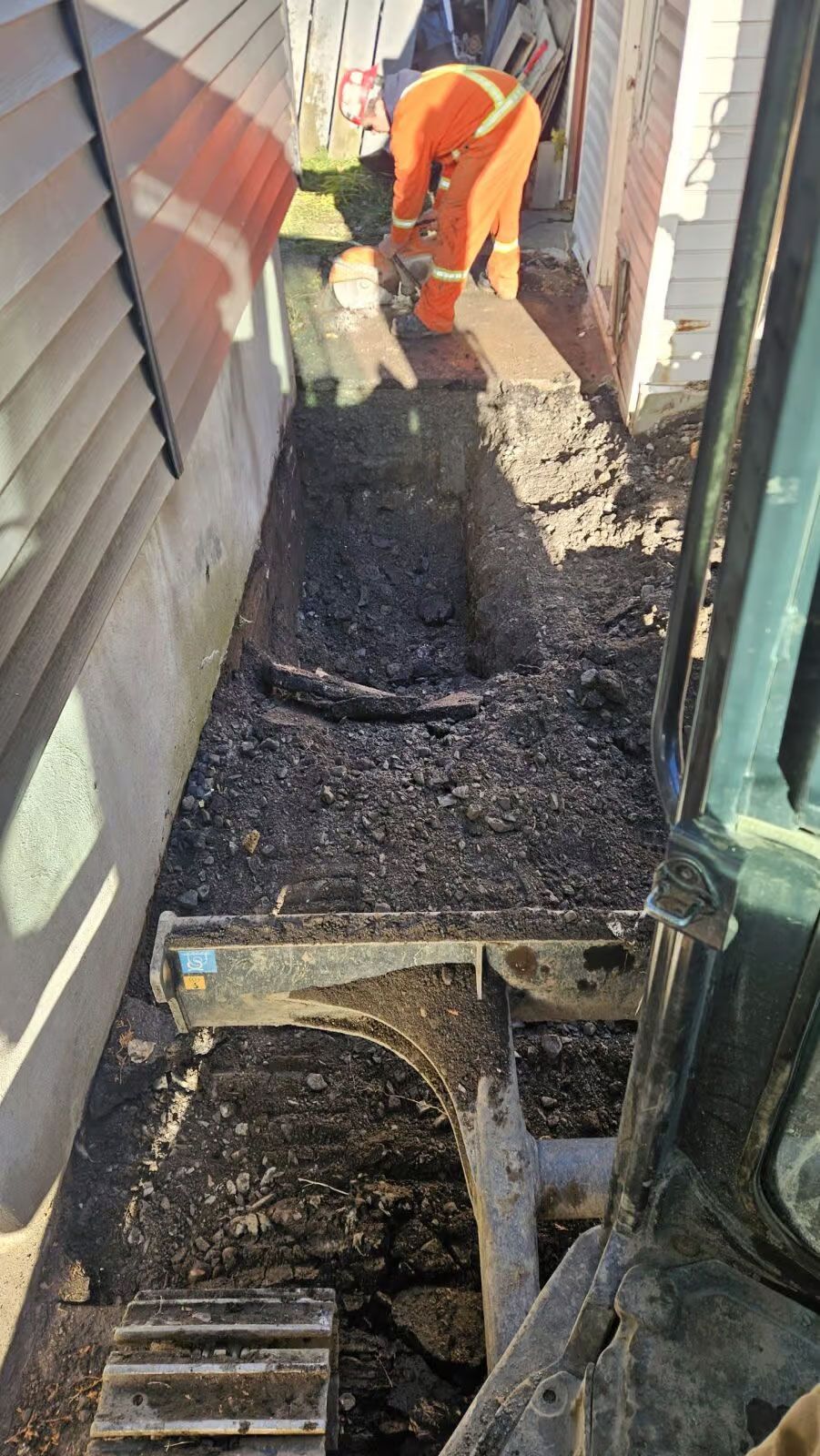 Un ouvrier en salopette orange répare une gouttière étroite sur un toit, recouverte d'asphalte humide et de débris.