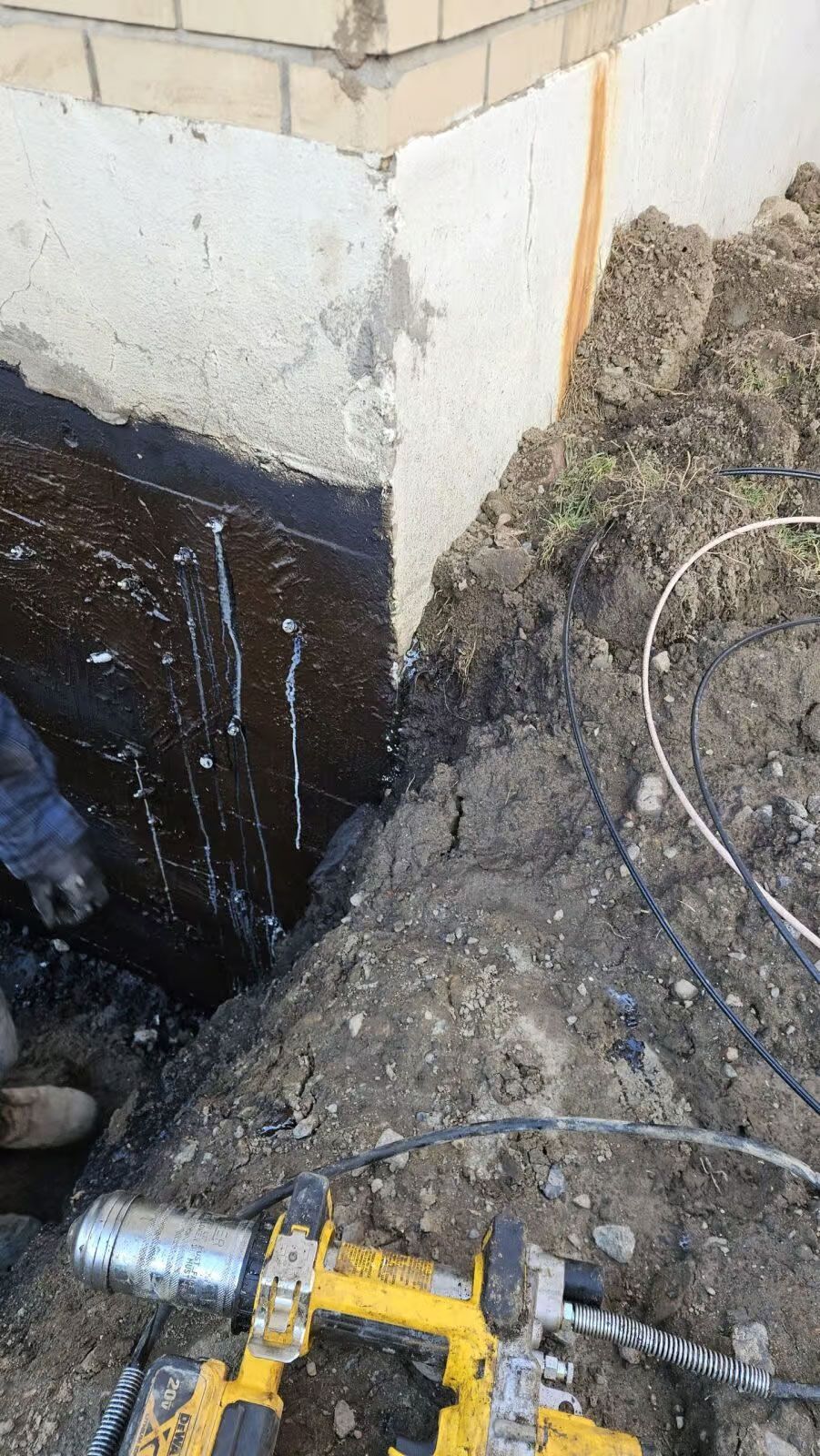 Réparation d'un coin de fondation ; enduit d'asphalte foncé sur béton, terre et outils jaunes visibles.