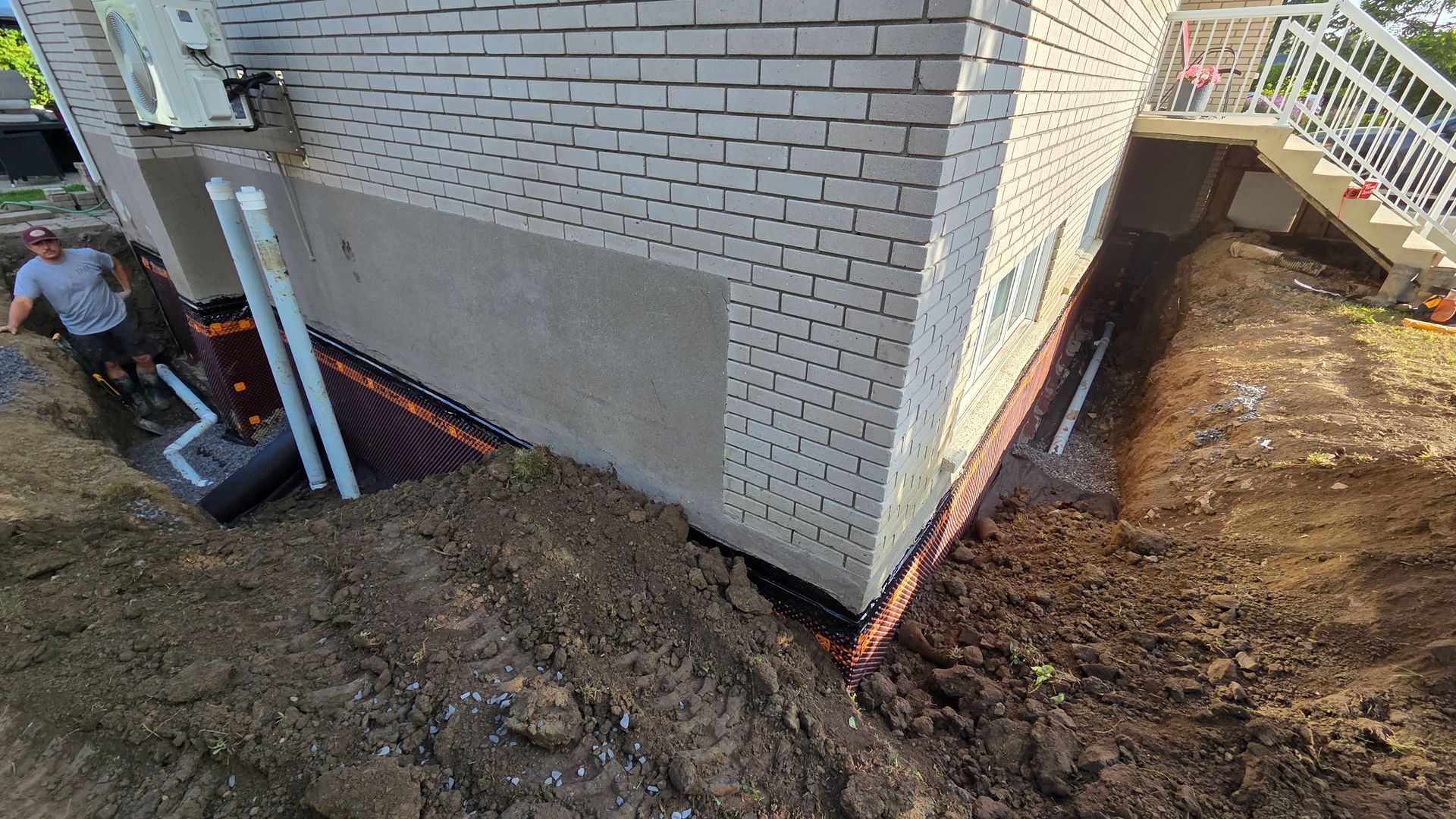 De la boue près des fondations d'une maison en briques lors de travaux de drainage ou de réparation extérieurs, avec des tuyaux et un ouvrier visibles.