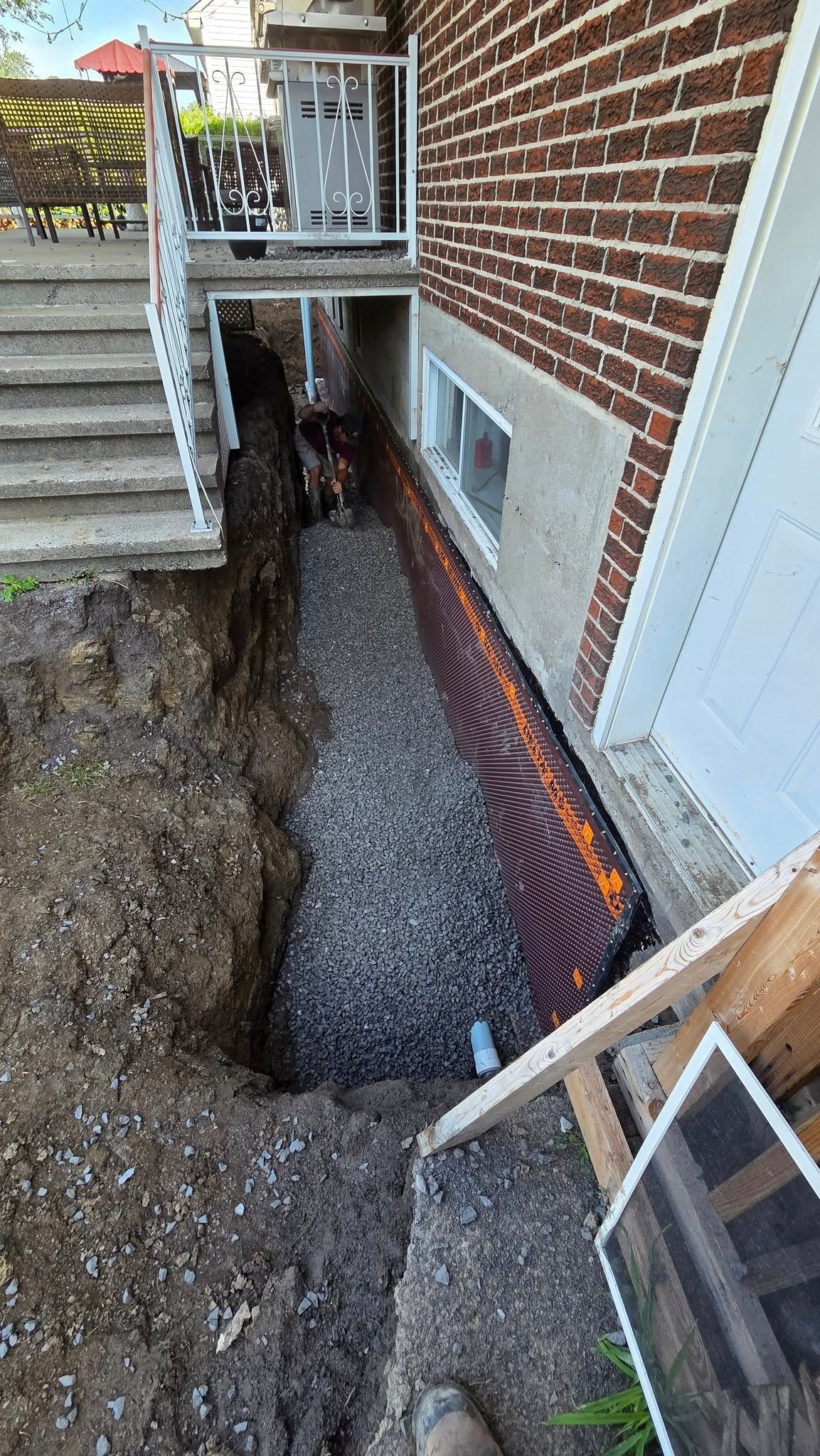 Tranchée étroite jouxtant une maison en briques, avec du gravier et des fondations apparentes pendant la construction.