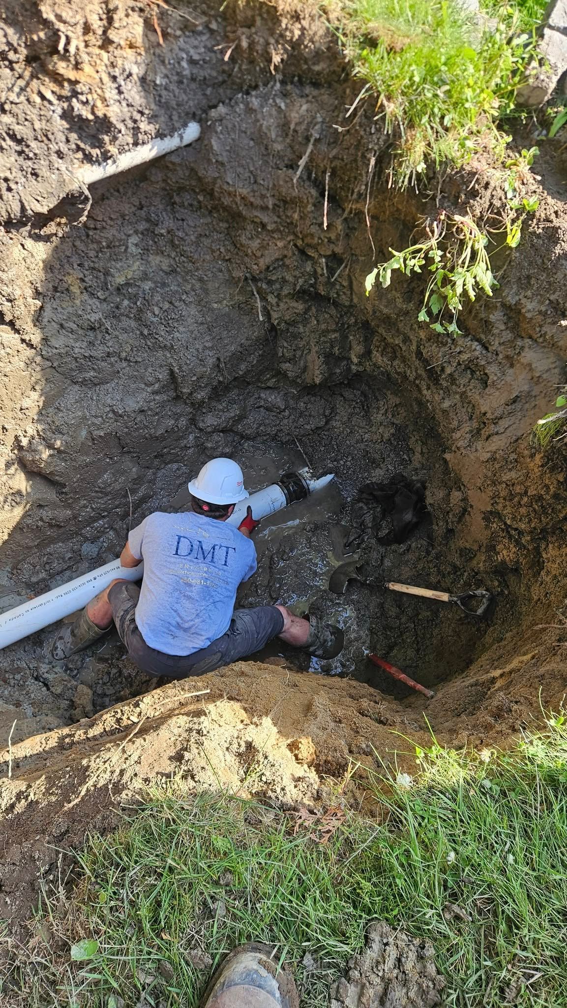 Un homme portant un casque de chantier travaille dans une tranchée profonde, raccordant des tuyaux blancs. À l'extérieur, près de l'herbe et de la terre.
