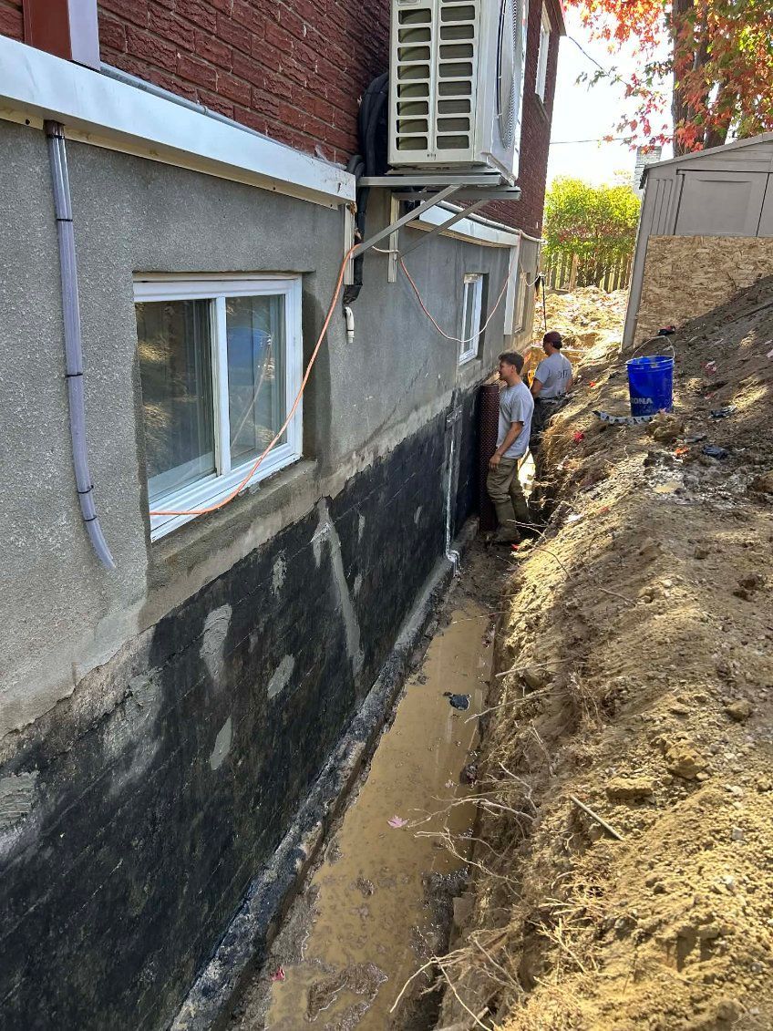 Des ouvriers effectuent des travaux d'excavation à proximité d'un bâtiment doté d'une membrane d'étanchéité noire nouvellement appliquée ; journée ensoleillée.