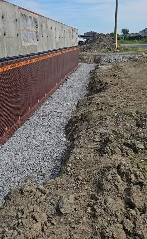 Fondation extérieure avec tranchée de drainage en gravier, membrane murale brune, chantier de construction.