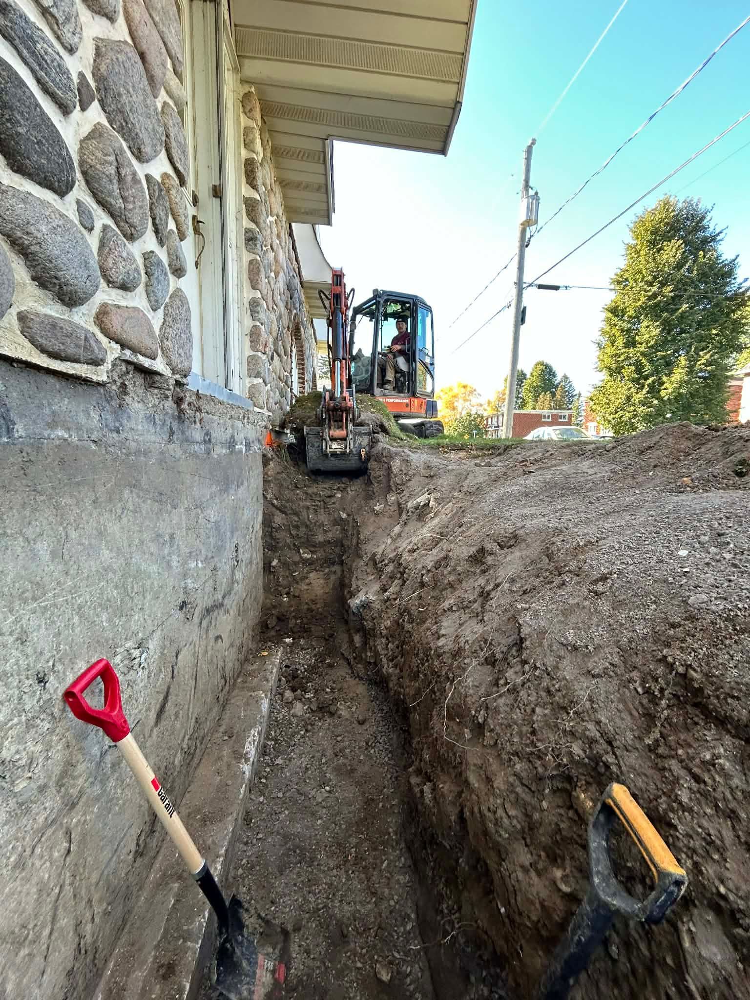 Une petite pelleteuse creuse une tranchée à côté d'un bâtiment en pierre. Des pelles sont appuyées contre des poteaux à proximité.