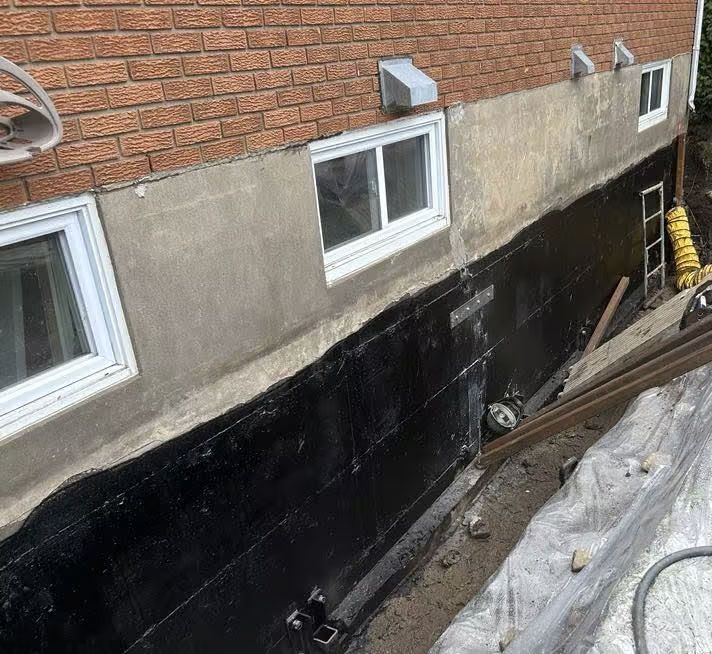 Mur de fondation en réparation à côté d'une maison en briques, avec des matériaux d'étanchéité et de construction noirs.