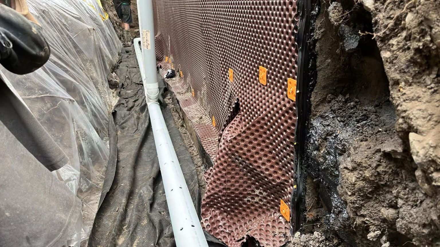 Tranchée étroite avec un tuyau blanc longeant une paroi rocheuse et une barrière en grillage couleur rouille.