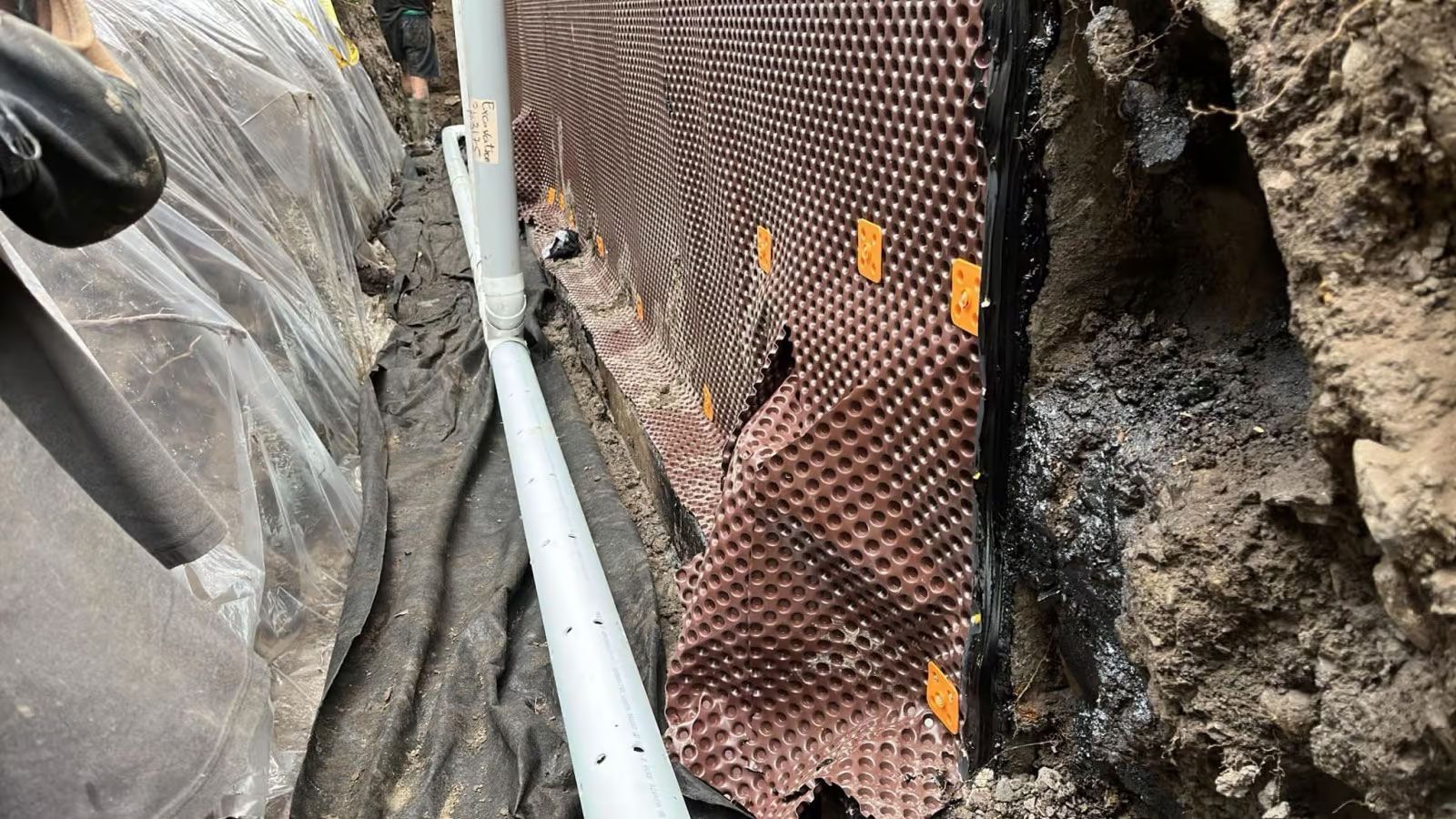 Système de drainage des fondations installé dans une tranchée. Tapis de drainage brun à picots contre le mur. Tuyau de drainage blanc.