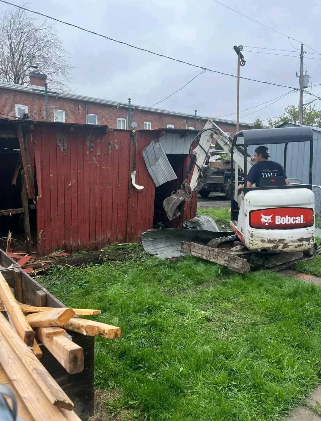 Une pelleteuse Bobcat démolit une structure en bois rouge. Une personne manœuvre l'engin sur une zone herbeuse.