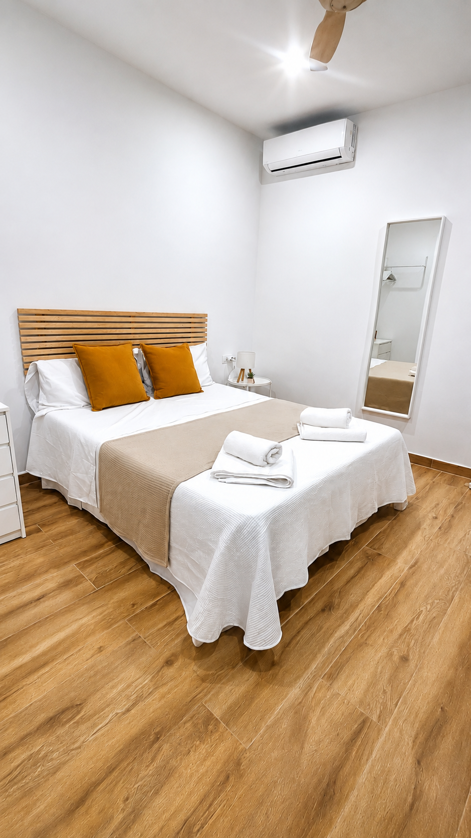 Habitación luminosa con cama, televisor montado en la pared, cómoda de madera, cortinas y cestas de mimbre.