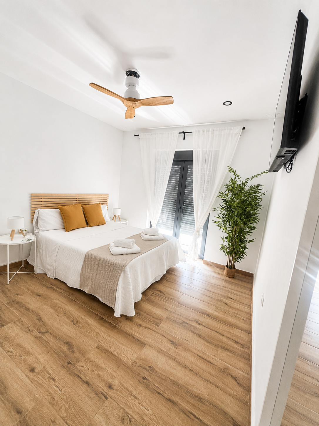 Habitación luminosa con cama, televisor montado en la pared, cómoda de madera, cortinas y cestas de mimbre.