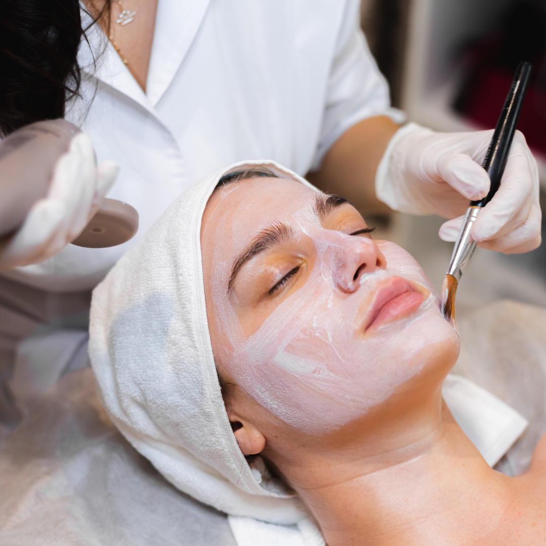 A professional wearing white gloves uses a brush to apply a white facial mask to a person lying down in a spa.