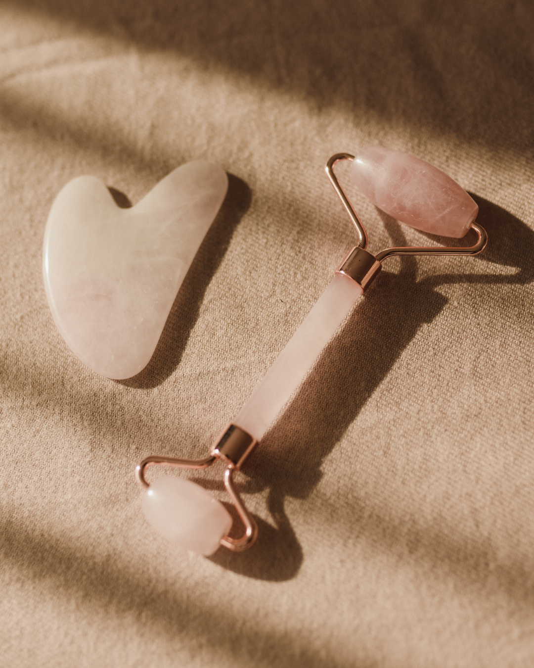 A rose quartz facial roller and a heart-shaped gua sha tool resting on a textured, light beige fabric.