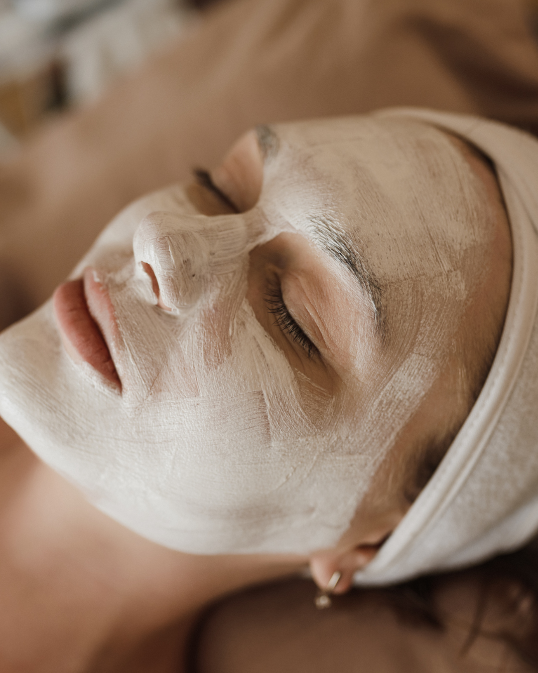 A person lying down with a dry, cracked beige clay face mask applied to their skin, eyes closed, wearing a white headband.