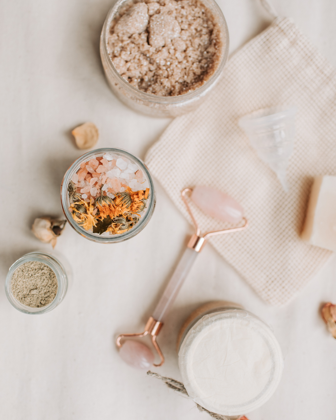 A flat lay of skincare products, including a rose quartz facial roller, jars of botanical salts, and a textured cloth bag.